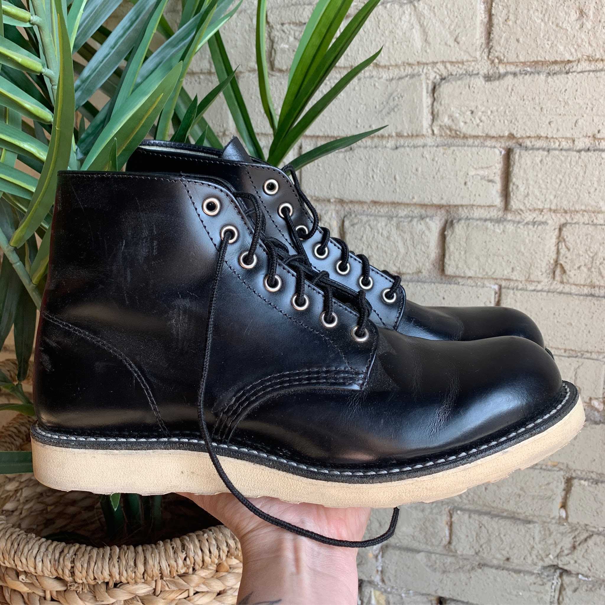 A pair of black leather lace-up work boots held up against white brick and a palm