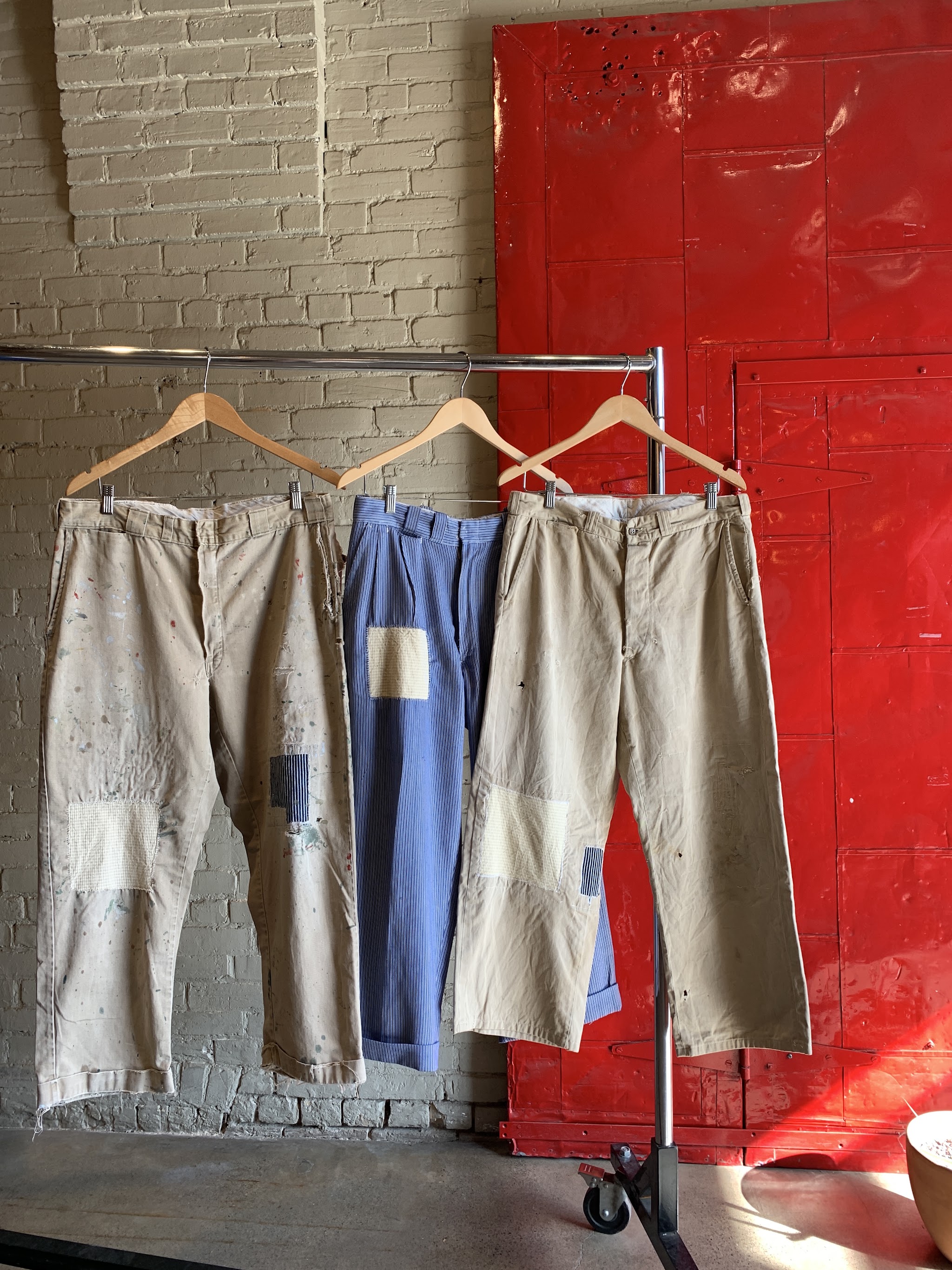 Three pairs of paint-splattered patched work pants on a rolling rack