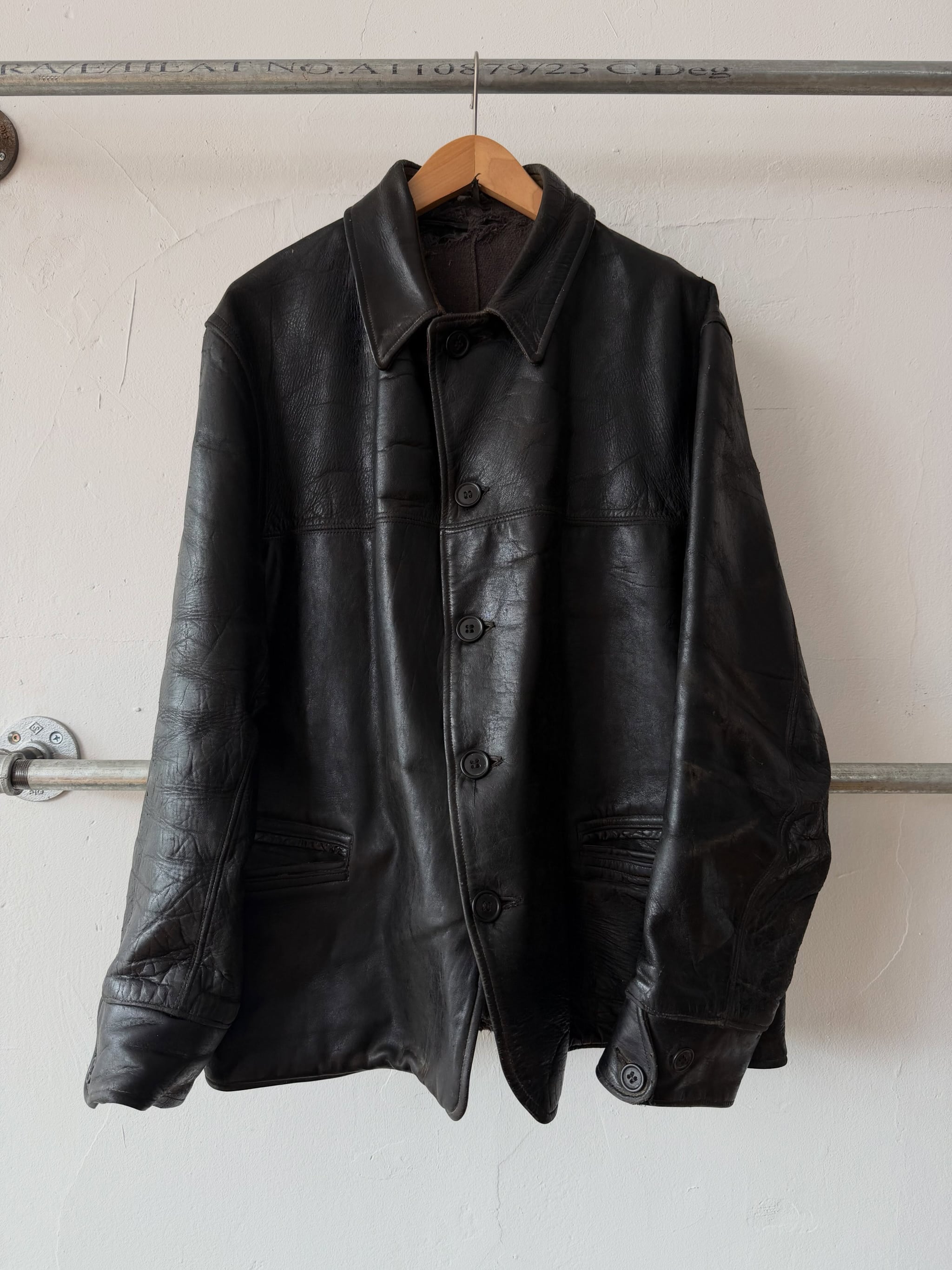 Black 1930s horsehide leather car coat hanging on a pipe rail against a white wall