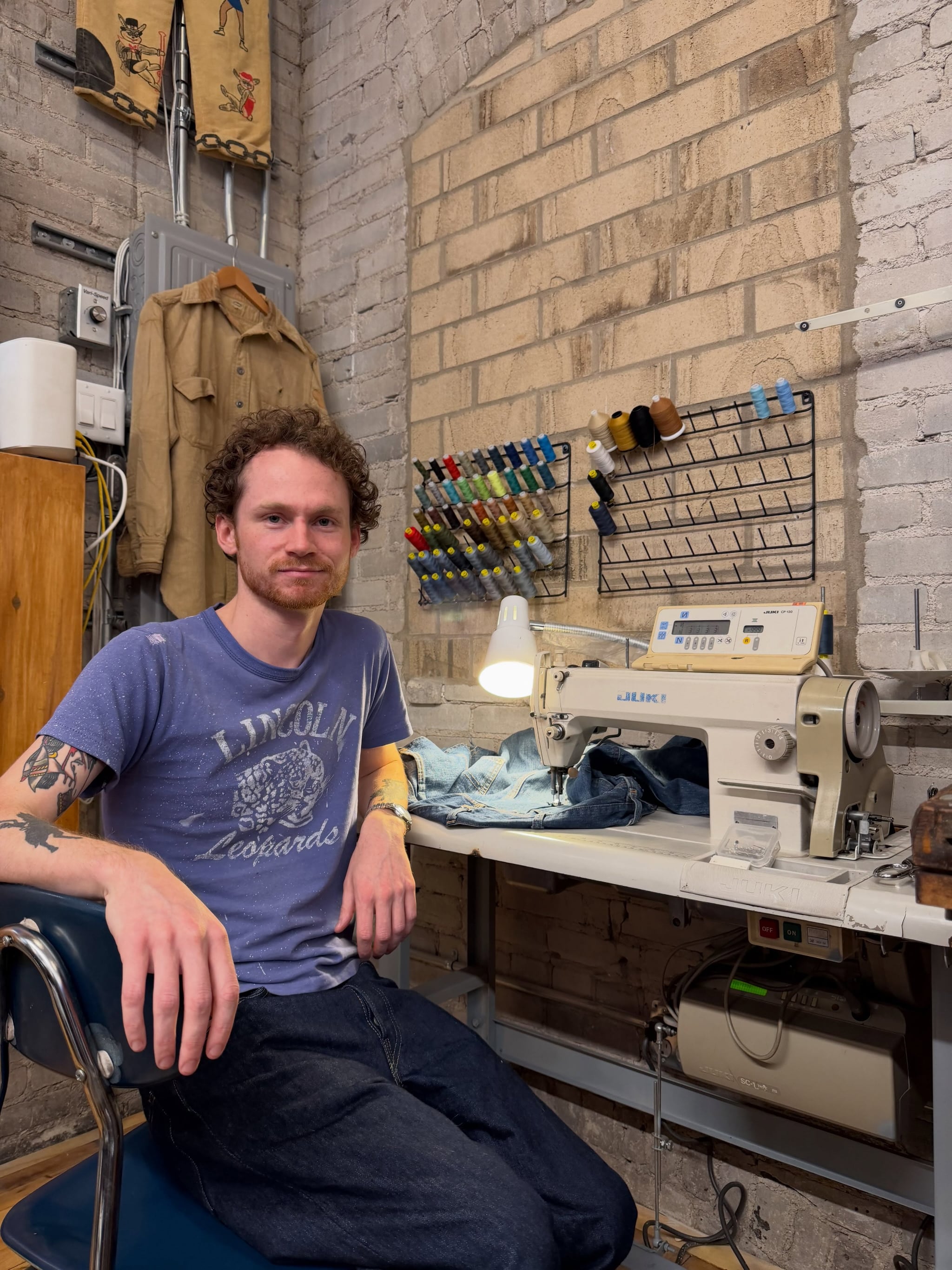 Izak, the shop’s tailor, sitting at an industrial sewing machine beside a wall of thread spools